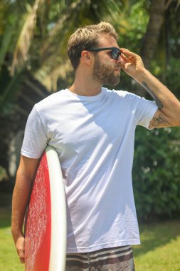 Bir adam canlı bir tropikal ortamda sörf tahtasıyla durur güneş gözlüğü takar ve heyecan verici bir maceraya hazırdır.