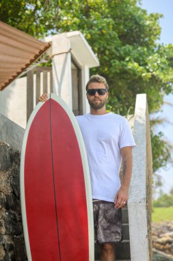 Şık bir sörfçü canlı kırmızı bir sörf tahtası tutuyor, güneşli bir kıyı şeridinde dalgalarda sörf yapmaya hazır.