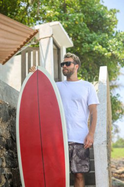 Şık bir sörfçü canlı kırmızı bir sörf tahtası tutuyor, güneşli bir kıyı şeridinde dalgalarda sörf yapmaya hazır.