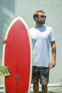 Şık bir sörfçü, güneşin altında yeni bir maceraya hevesle hazır canlı bir kırmızı sörf tahtasıyla kendine güvenerek duruyor.