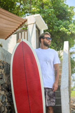 Günlük plaj kıyafeti giymiş bir adam parlak renkli bir sörf tahtasının yanında duruyor ve yaz havasını tamamen kucaklıyor.