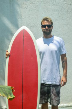 Şık bir sörfçü, güneşin altında yeni bir maceraya hevesle hazır canlı bir kırmızı sörf tahtasıyla kendine güvenerek duruyor.