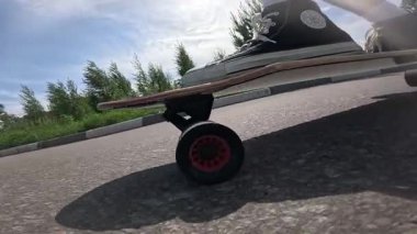 Güneşli bir gökyüzünün altında asfalt bir yolda spor ayakkabılarla uzun bir sörf tahtası süren birinin yavaş çekim görüntüleri. Düşük açıdan çekilen video, bir yaz günü boyunca hareket, yaşam tarzı ve özgürlüğü vurguluyor..