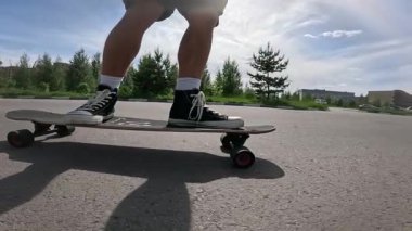 Güneşli bir gökyüzünün altında asfalt bir yolda spor ayakkabılarla uzun bir sörf tahtası süren birinin yavaş çekim görüntüleri. Düşük açıdan çekilen video, bir yaz günü boyunca hareket, yaşam tarzı ve özgürlüğü vurguluyor..