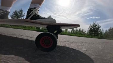 Güneşli bir gökyüzünün altında asfalt bir yolda spor ayakkabılarla uzun bir sörf tahtası süren birinin yavaş çekim görüntüleri. Düşük açıdan çekilen video, bir yaz günü boyunca hareket, yaşam tarzı ve özgürlüğü vurguluyor..