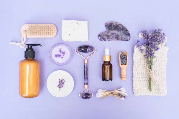 Lavender cosmetics for skin care - cosmetic oil, handmade soap, bath salt, body lotion with lavender extract and gua sha massager over purple background. Flat lay style