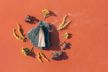 Yanan palo santo çubuğu, turuncu kırmızı arka planda güzel kokulu çiçekler ve otlarla tütüyor. Meditasyon ve ruhani uygulamalar için kutsal tahta çubuklar. Oda dezenfekte ayini. Seçici odak