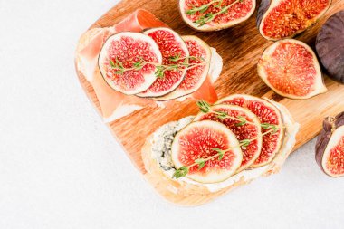 Üzerinde çiğ incir, peynir, prosciutto ve otlar olan taze bruschetaları tahta kesme tahtasının üzerinde fotokopi alanı ile birlikte beyaz beton masa zemininin üzerinde görebilirsiniz. Sağlıklı bir kahvaltı.