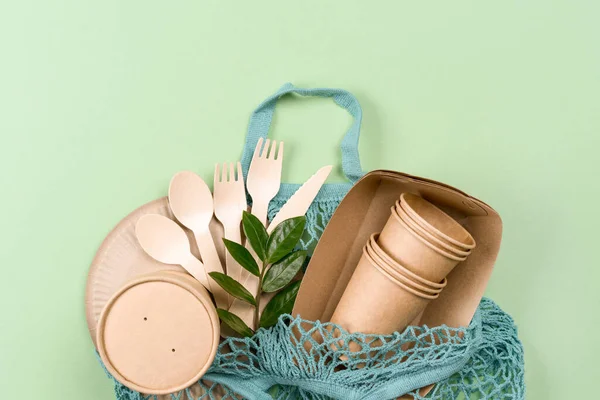 Paper utensils in blue cotton bag over light green background. Kraft paper cups, food containers and bamboo cutlery set - paper tableware. Sustainable food packaging concept
