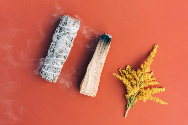 Burning incense - white sage and palo santo stick with smoke over orange brown background. Bundle for meditation and room fumigation. Selective focus. Flat lay style