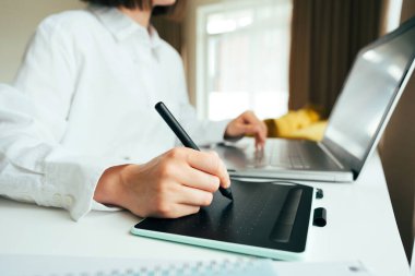 Modern iç mekanda dizüstü bilgisayar ve grafik tablet kullanan genç kadın grafik tasarımcı veya dijital ressamın yüzsüz görüntüsü. Ev ofisinden uzaktan çalışan serbest çalışan biri. Yakın görünüm