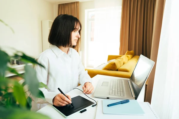 Caucasian young woman graphic designer or digital artist using laptop computer and graphic tablet in the modern home office interior. Freelancer working remotely from home