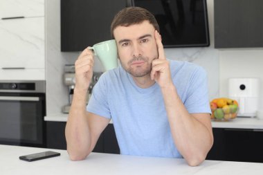 Mutfakta baş ağrısı sırasında kafasının yanında bir fincan kahve tutan yakışıklı genç adamın portresi.