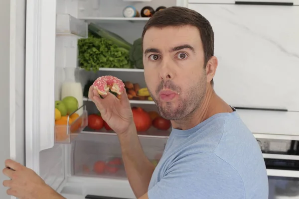 一个英俊的年轻人在厨房吃冰箱里的甜甜圈的肖像 — 图库照片