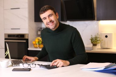 Mutfakta dizüstü bilgisayarla çalışan yakışıklı bir gencin portresi.