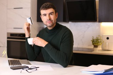 Mutfakta kredi kartı ve dizüstü bilgisayarla e-alışveriş yapan yakışıklı genç adamın portresi.