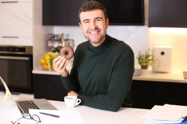 Mutfakta çalışırken kahve eşliğinde çörek yiyen yakışıklı bir gencin portresi.