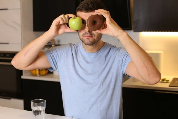 健康的なリンゴと不健康なドーナツを持っているハンサムな若い男の肖像画彼の目の前でキッチン — ストック写真