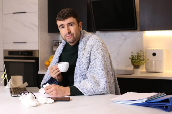 portrait of handsome young ill man with blanket working from home at kitchen