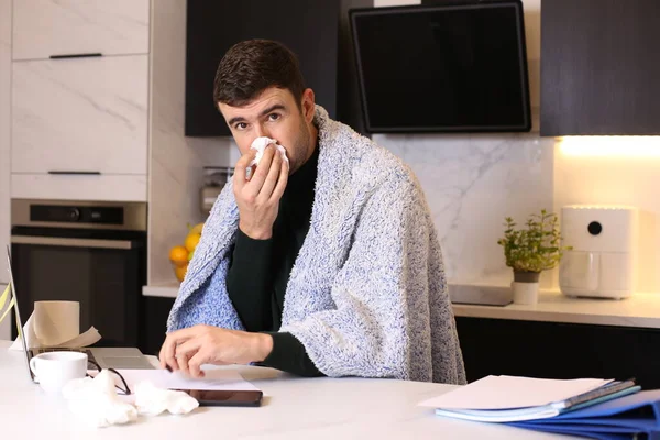 portrait of handsome young ill man with blanket working from home at kitchen