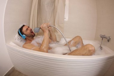 wide angle shot of handsome young man with headphones singing with shower head in bath