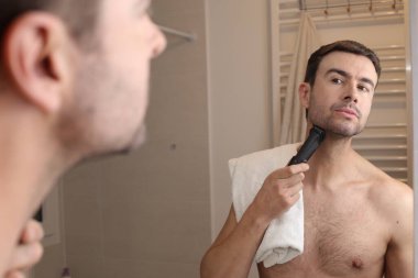 Banyoda aynanın önünde sakalını elektrikli makasla tıraş eden yakışıklı genç adamın portresi.