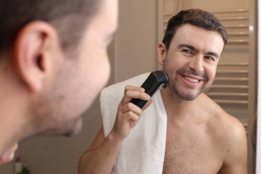 Banyoda aynanın önünde sakalını elektrikli tıraş makinesiyle tıraş eden yakışıklı genç adamın portresi.