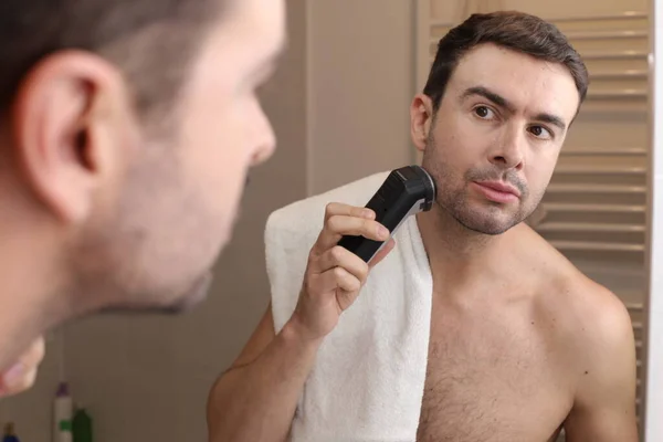 浴室の鏡の前で電気シェーバーで髭を剃るハンサムな若い男の肖像画 — ストック写真