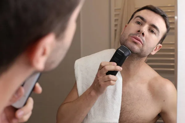 浴室の鏡の前で電気シェーバーで髭を剃るハンサムな若い男の肖像画 — ストック写真