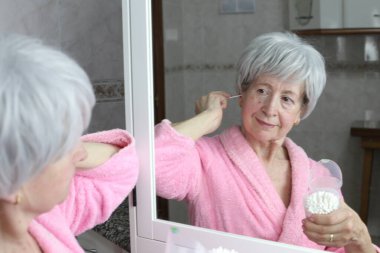 Banyoda aynanın önünde pamuklu çubuklarla kulakları temizleyen olgun bir kadının yakın plan portresi.