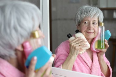 Banyoda aynanın önünde elinde şişe cilt losyonu olan olgun bir kadının yakın plan portresi.