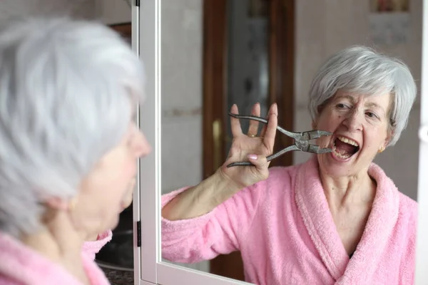 close-up portrait of mature woman pulling out her tooth with pliers in front of mirror in bathroom