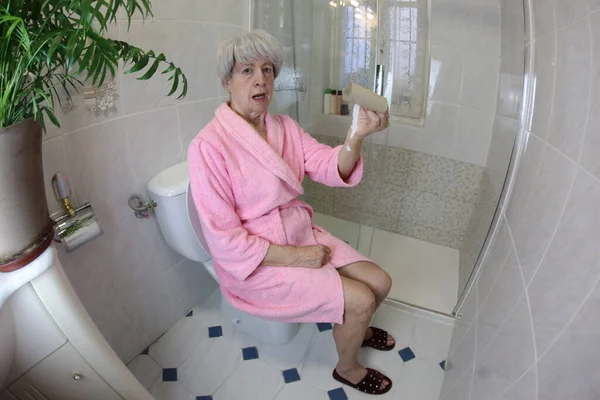 トイレに座っている空のトイレットペーパーのロールを持つシニア女性のワイドアングルショット — ストック写真