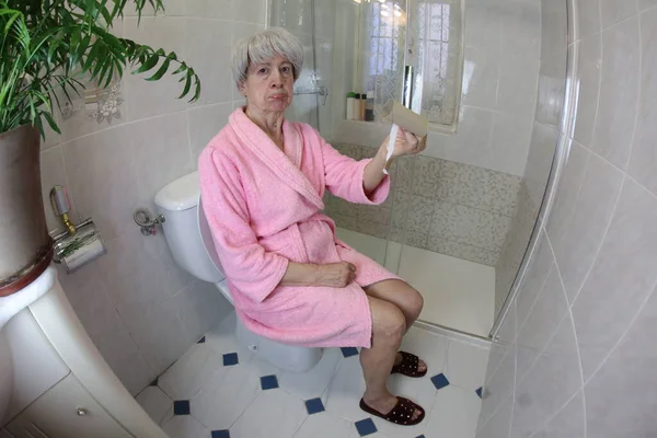 トイレに座っている空のトイレットペーパーのロールを持つシニア女性のワイドアングルショット — ストック写真
