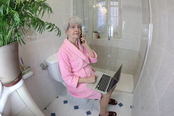 wide angle shot of senior woman talking by phone and working with laptop in toilet