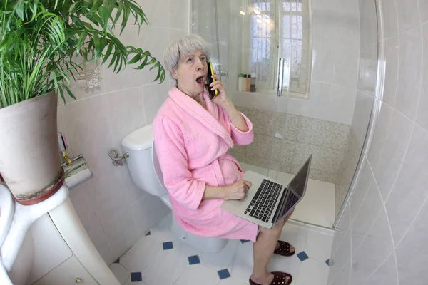 wide angle shot of senior woman talking by phone and working with laptop in toilet