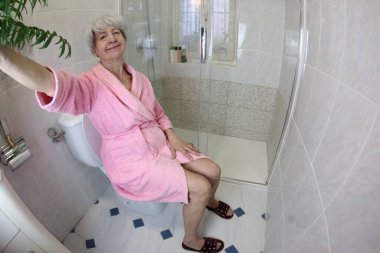 Banyoda tuvalette otururken selfie çeken yaşlı bir kadının geniş açılı görüntüsü.