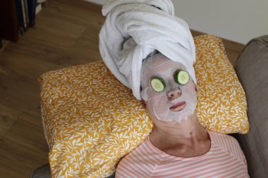 Kafasında havluyla rahatlayan olgun bir kadının yakın plan portresi, yüz maskesi ve gözlerinde salatalık.