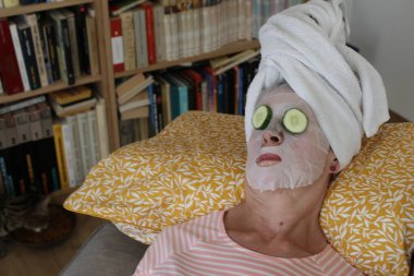 Kafasında havluyla rahatlayan olgun bir kadının yakın plan portresi, yüz maskesi ve gözlerinde salatalık.