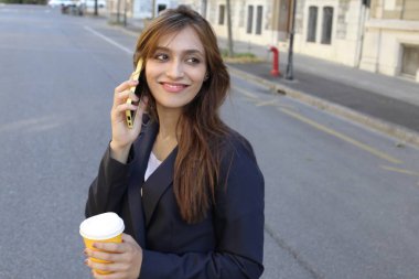 Takım elbiseli iş kadını akıllı telefonla konuşuyor ve dışarıda kahve tutuyor.