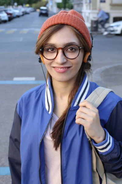 戴着便帽和眼镜的女学生通过耳机在户外听音乐 — 图库照片