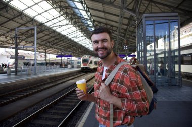Sırt çantasıyla seyahat ederken tren istasyonunda bekleyen yakışıklı, mutlu genç adamın portresi.