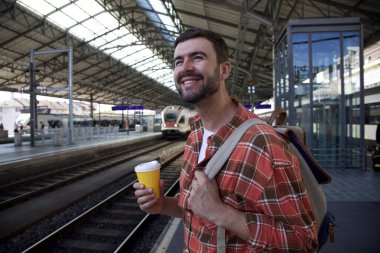 Sırt çantasıyla seyahat ederken tren istasyonunda bekleyen yakışıklı, mutlu genç adamın portresi.