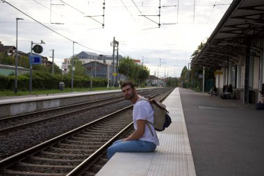 Sırt çantasıyla seyahat ederken tren istasyonunda bekleyen yakışıklı, mutlu genç adamın portresi.