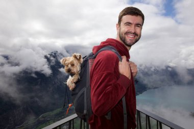 Sırt çantasında sevimli köpeğiyle gezen yakışıklı, mutlu genç adamın portresi.