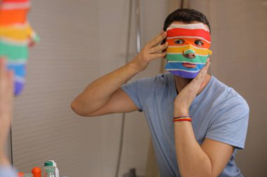 Yüzü maskeli yakışıklı genç bir adamın portresi. Banyoda aynanın önünde lgbtqa bayrağı renginde.