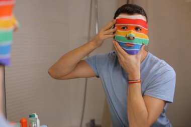 Yüzü maskeli yakışıklı genç bir adamın portresi. Banyoda aynanın önünde lgbtqa bayrağı renginde.