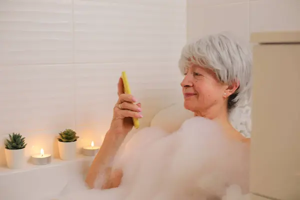 快乐的老年妇女的肖像 她在浴缸里拿着智能手机 还有肥皂泡 — 图库照片