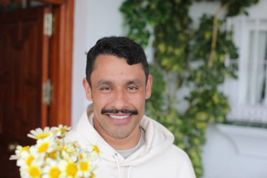 Beyaz kapüşonlu bıyıklı yakışıklı İspanyol adamın portresi gülümsüyor ve kameraya papatya çiçeği veriyor.