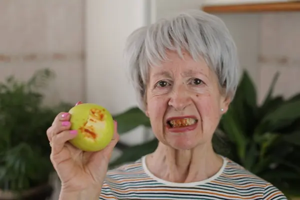 自宅で緑のリンゴを食べる出血歯を持つ成熟した灰色の髪の女性の肖像画 — ストック写真